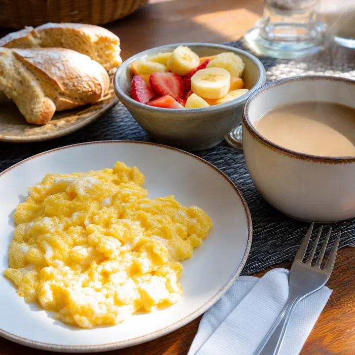 An Argentine breakfast table with a plate of scrambled eggs, cut fresh fruit, bread and a steaming cup of coffee with milk, under the morning light coming through the window.