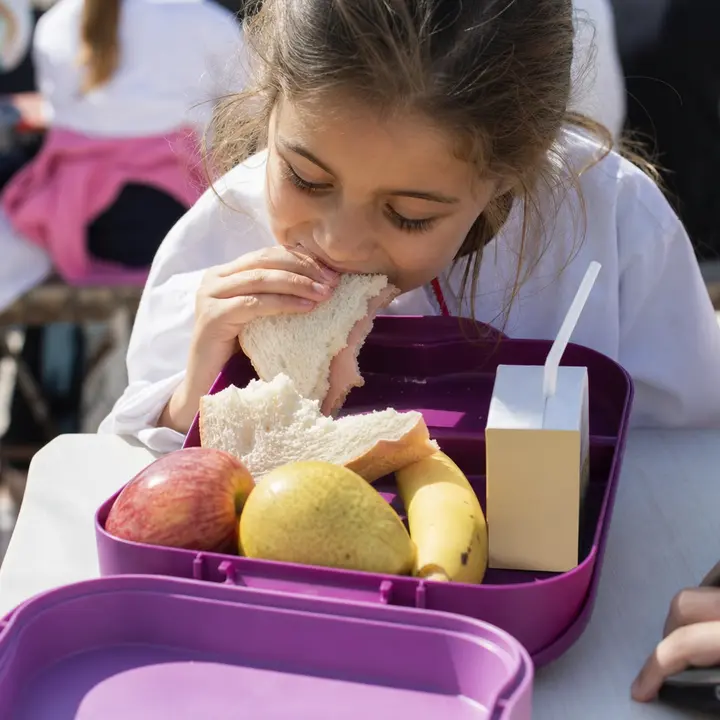 A girl in a school uniform bites into a sandwich while sitting in front of an open lunchbox containing an apple, a pear, a banana, and a juice box.