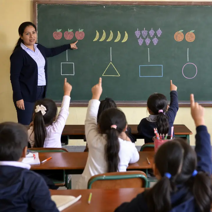 A teacher at a chalkboard points at drawn groups of objects and shapes while seated children watch and raise their hands.