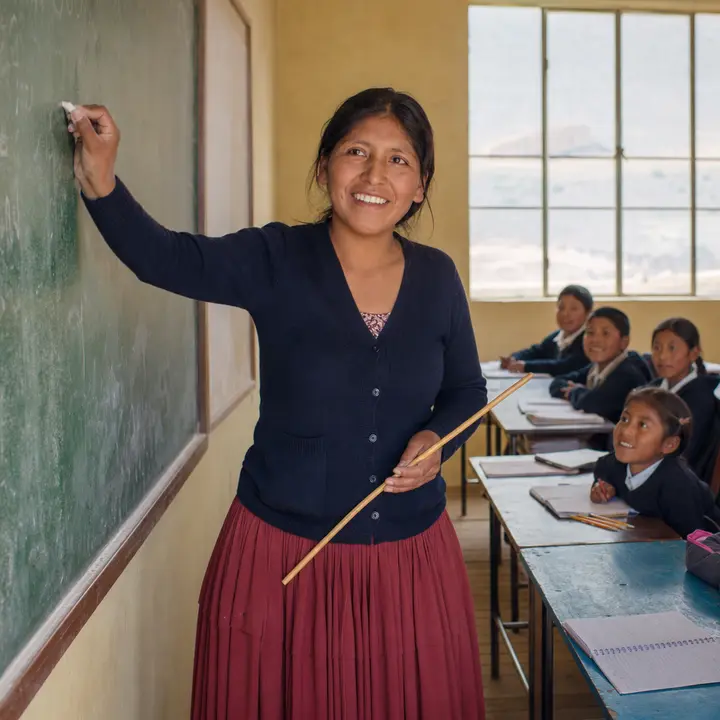 A smiling Bolivian teacher, standing in front of a chalkboard, writes with chalk while holding a pointer and looking at her class.